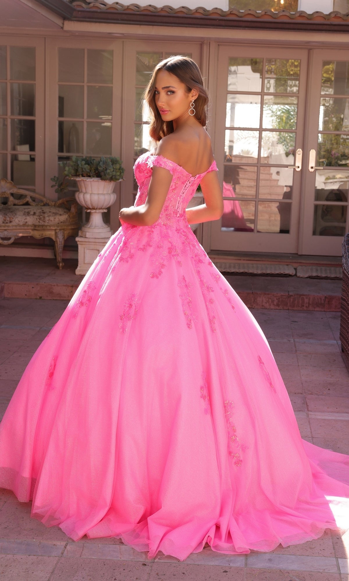 A woman stands outdoors in front of glass doors, wearing the Off-the-Shoulder Lace-Up Long Prom Ball Gown H1352—a bright pink gown with floral details and a full skirt.